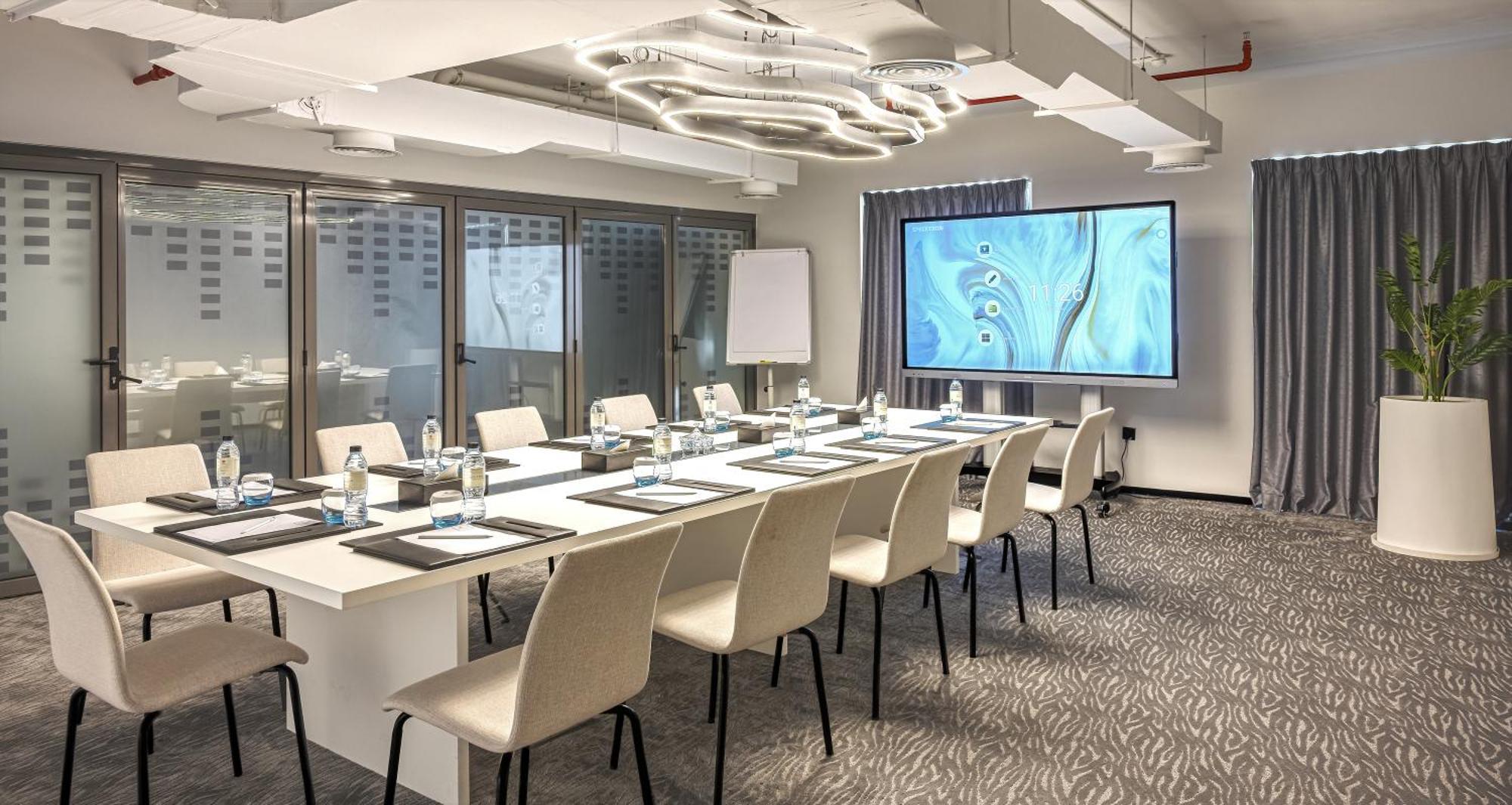 Millennium Atria Business Bay Aparthotel Dubai Bagian luar foto The photo shows a modern conference room set up for a meeting. There is a long table with several chairs arranged around it, each with a notepad and pen. A large screen displays a graphic or presentation, and there is a whiteboard in the corner. The 