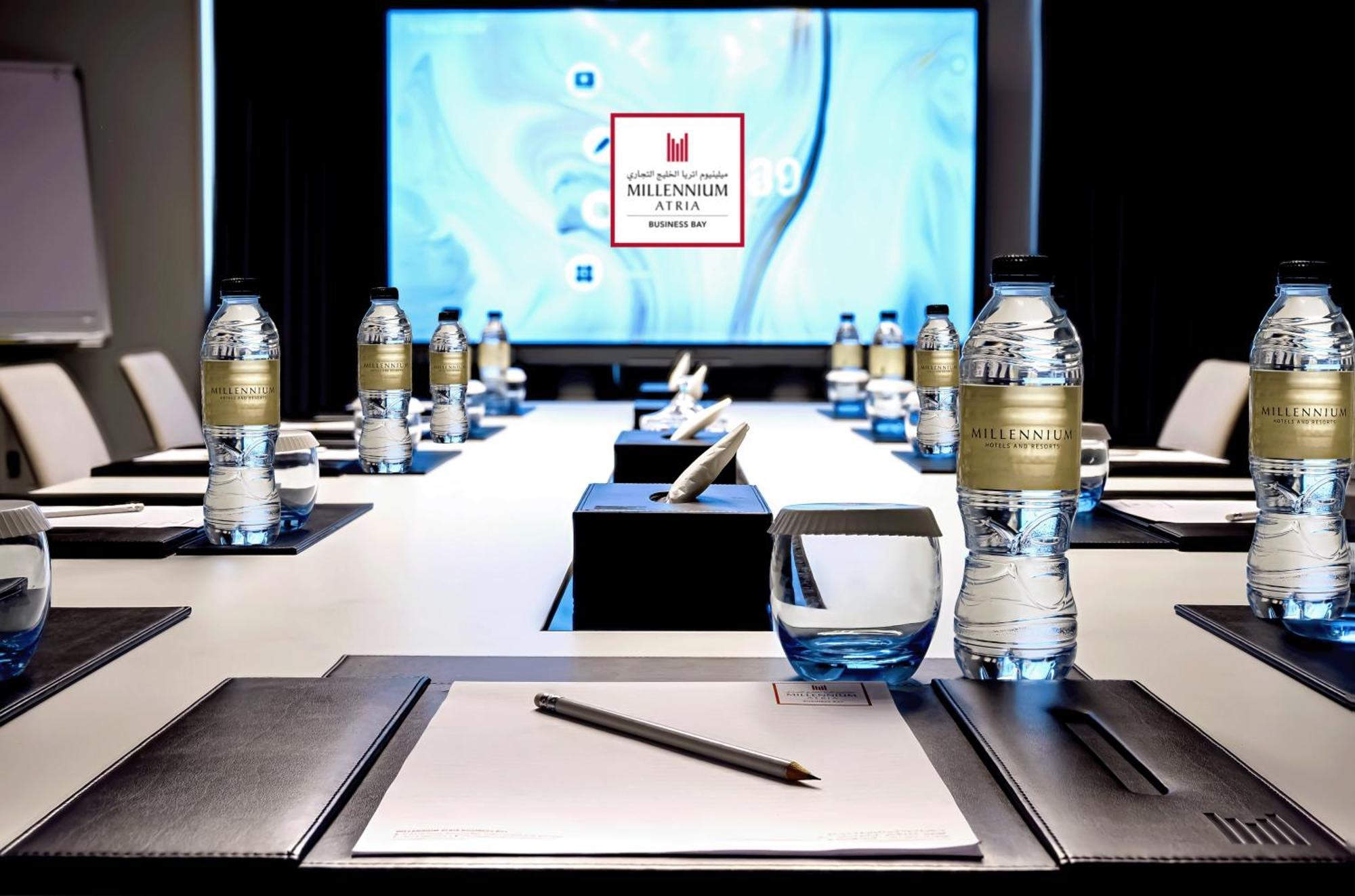 Millennium Atria Business Bay Aparthotel Dubai Bagian luar foto The photo shows a modern conference room setup. A long table is laid out with neatly arranged stationery, including notebooks and pens, at each seat. Several bottles of water are placed on the table. In the background, a large screen is displayed, li