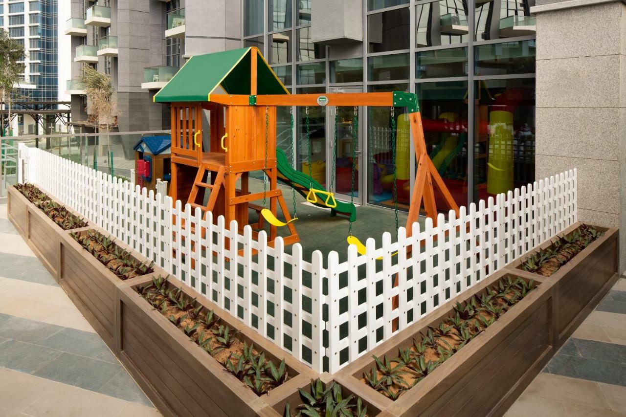 Millennium Atria Business Bay Aparthotel Dubai Bagian luar foto The image shows a children's playground area, which is enclosed by a white picket fence. The playground features a wooden play structure with a slide, swings, and climbing elements. Surrounding the play area are planters with greenery, and the backdr
