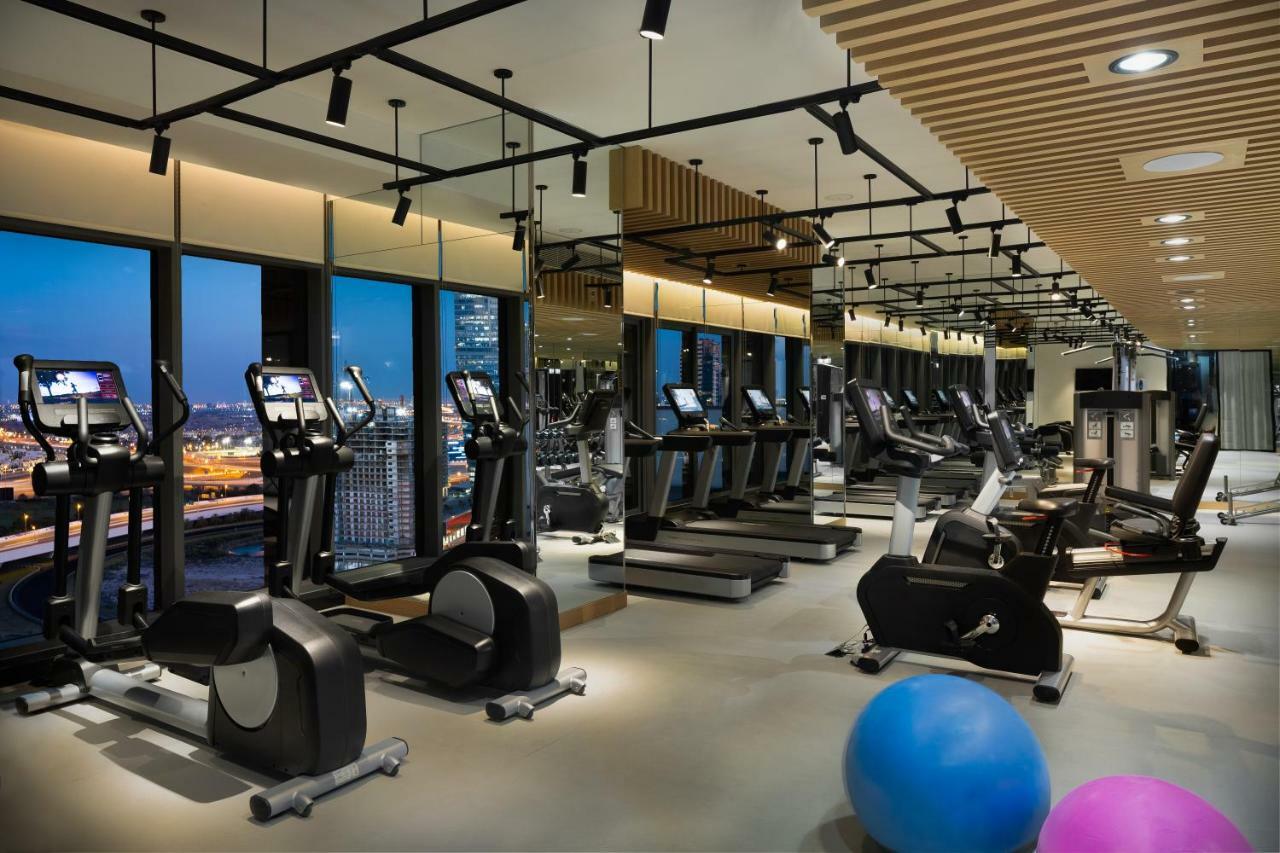 Millennium Atria Business Bay Aparthotel Dubai Bagian luar foto The photo shows a modern gym with various types of exercise equipment available. There are several treadmills and elliptical machines arranged in rows, with large windows in the background providing a view of the city. The lighting is bright and cont