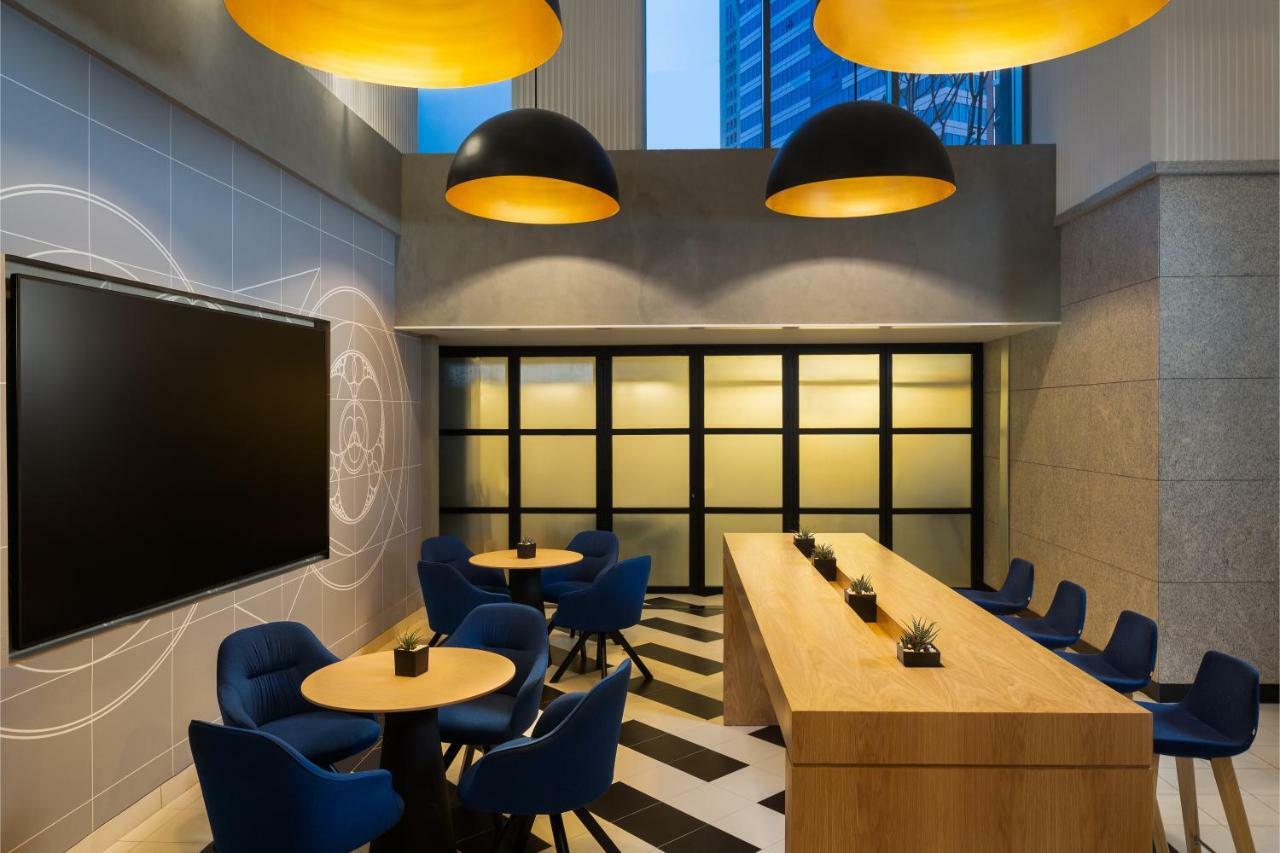 Millennium Atria Business Bay Aparthotel Dubai Bagian luar foto The photo shows a modern interior space, likely a lounge or meeting area. It features a long wooden table with some small plants on it, accompanied by several blue, circular chairs arranged around it. There is a large flat-screen television mounted o