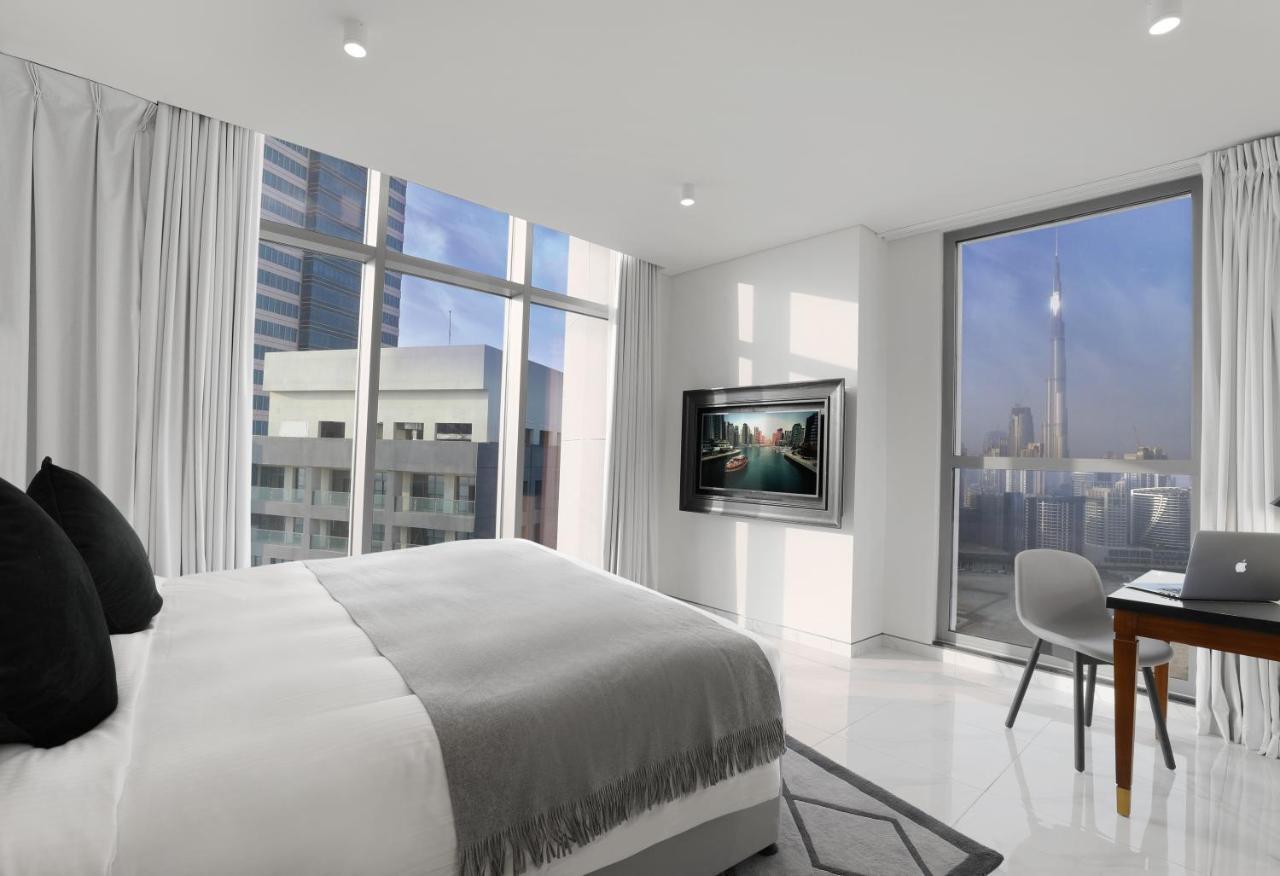Millennium Atria Business Bay Aparthotel Dubai Bagian luar foto The photo depicts a modern hotel room that features a large bed with a gray throw blanket. There are large windows allowing natural light to fill the space, showcasing a city skyline view. A flat-screen television is mounted on the wall opposite the 